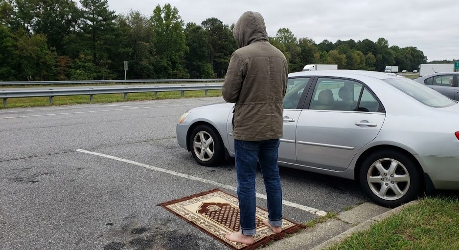 Une personne se préparant à la prière sur un tapis de voyage près d'une voiture sur une aire d'autoroute, illustrant le besoin de solutions de wudu portables.